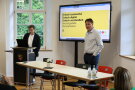 Markus Donhauser und Fabian Blomeyer sprechen zu den Gästen, im Hintergrund eine Präsentation