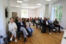 Gäste sitzen auf Stühlen in einem Raum und hören dem Redner zu