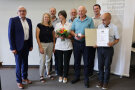 Eine Gruppe von Menschen. Einer hält eine Schatulle mit Medaille, einer eine Urkunde und eine Frau einen Blumenstrauß