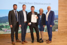 Fünf Personen nebeneinander. Mittig hält ein Mann eine Medaille und Urkunde.