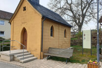 Eine Kapelle mit einer Sitzbank davor und einer Informationstafel