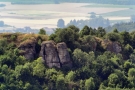 Blick auf eine hoch aufragende mit Bäumen und Sträuchern bewachsene Feldformation und eine flache Landschaft mit Dorf im Hintergrund. 