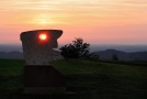 Aus einer Steinplatte herausgearbeiteter stilisierter menschlicher Kopf steht auf einem Aussichtspunkt in einer Landschaft. Die untergehende Sonne leuchtet genau durch das Loch in der Steinplatte, mit dem der Künstler das Auge dargestellt hat.