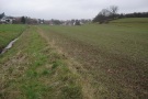 Ein kerzengerader Bachlauf mit einem Acker auf der einen Uferseite, der bis an die Böschungskante reicht. Im Hintergrund eine Ortschaft.