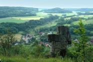 Blick von einem Hügel, auf dem eine kreuzartige Felsformation steht, auf ein Dorf mit einer reich strukturierten Landschaft im Hintergrund.