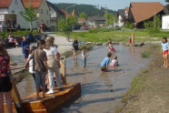 Kinder beim Spielen in einem flachen Bach, der mit einem kleinen Floss ausgestattet und zum Wasserspielplatz umgestaltet wurde.