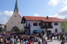 Fest auf dem Dorfplatz mit vielen Menschen. Rechts ein zweigeschossiges Wohnhaus (typisch oberbayerisches Satteldach mit geringer Dachneigung). Links dahinter Kirche.