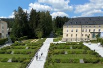 In der großen Parkgrünfläche verlaufen Fußwege und quer dazu Buschpflanzungen. Der Park ist von hohen Bäumen und cremefarbenen Kurgebäuden eingefasst.